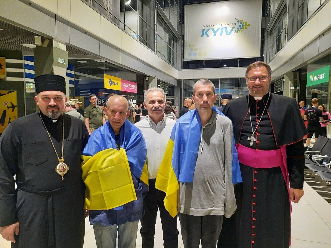 Draped in Ukrainian flags, Redemptorist Fathers Bohdan Geleta, left, and Ivan Levytsky, right, pose for a photo at the Kyiv airport in Ukraine June 29, 2024. To the far right is Archbishop Visvaldas Kulbokas, apostolic nuncio to Ukraine. (CNS photo/Courtesy Ukrainian Greek Catholic Church Communications Department)