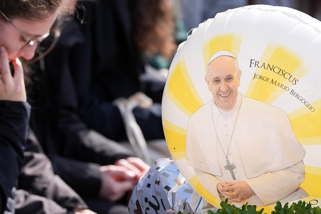 A balloon featuring an image of Pope Francis is seen as people pray around a statue of St. John Paul II outside Rome’s Gemelli hospital March 2, 2025. Pope Francis is receiving treatment there for double pneumonia. (CNS photo/Lola Gomez)